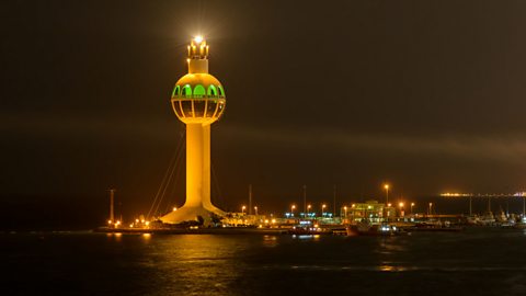 A tall tower, with a dome two thirds up, lights up the night sky around Jeddah's Seaport in Saudi Arabia. There is a beacon shining on top.