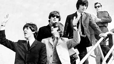 A black and white photo of Brian Epstein stood behind The Beatles as they get off a plane (Credit: Getty Images)