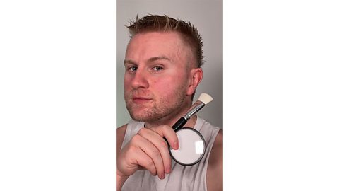 Man with a clean face holds a makeup brush and white face paint