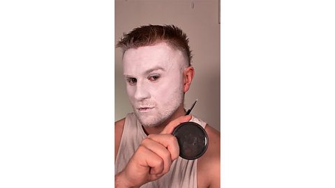 Man with painted white face holds a small makeup brush and black face paint