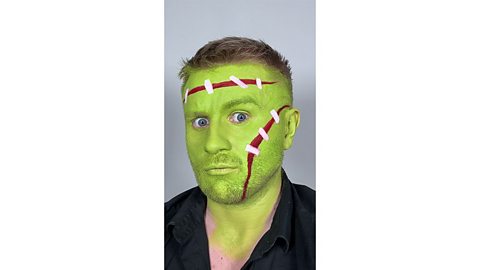 Man's face is painted green with red stripes on their forehead and cheek to look like scars