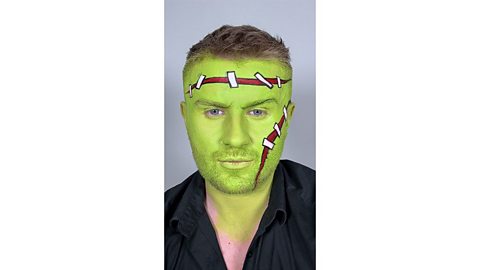 Man's face is painted green with fake scars painted on forehead and cheek, added detail with white and black stitches