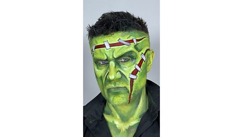 Man's face is painted green with fake scars painted on forehead and cheek, added detail with grey-green shading to age them plus white and black stitches