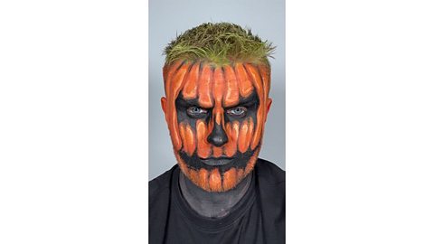 Man with orange painted face, black shadows around the eyes and mouth and added dark and light shadows and highlights to make them look pumpkin-like. Added green tint to their hair and dark coloured neck