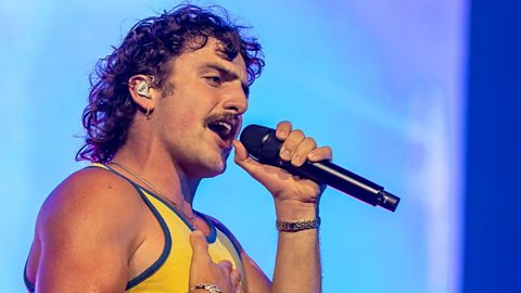 Benson Boone sings into a microphone wearing a yellow vest, his hair in a curly mullet and moustache. Bright blue background