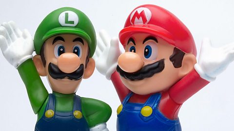 Toy models of video game characters Mario and Luigi wearing their blue dungarees, Luigi with green top and hat then Mario in red top and hat. Both with thick dark moustaches