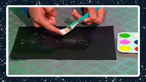 A pair of hands flicking white paint onto some black paint with a palette of paint next to it.