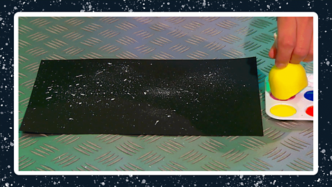 A piece of black card with white paint flicked onto it, next to a hand dabbing a sponge into some red paint