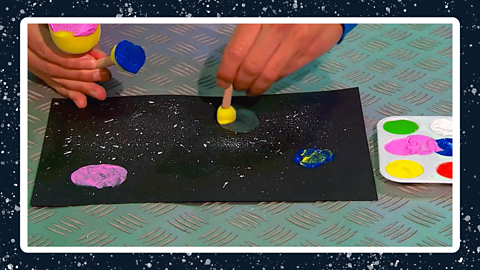 A pair of hands dabbing sponges into black card in different colours next to a paint palette