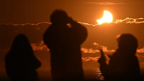 The silhouettes of three people watching the beginnings of a solar eclipse above the ocean (Credit: Getty Images)