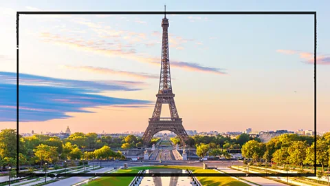 The Eiffel Tower in Paris is shown against a blue sky with orange light at the horizon. The sky is full of wispy white and grey clouds and the tower is surrounded by trees illuminated by golden light (Credit: Getty Images)
