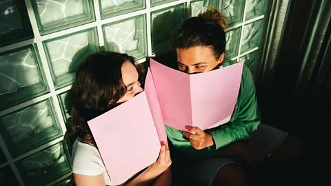 Two friends smile at each other from behind pink menus (Credit: Getty Images)
