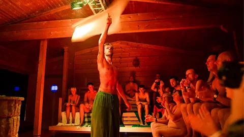 An Aufgussmeister twirls a towel in a sauna as a crowd watches around him (Credit: Getty Images)