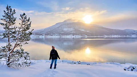 Person standing on a snowy shoreline overlooking a calm lake at sunrise with snow-covered mountain reflected in water (Credit: Alamy)