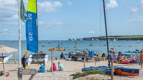 A busy summer sandy beach, Studland Bay, with various watersports going on and sail boats further out in the water.
