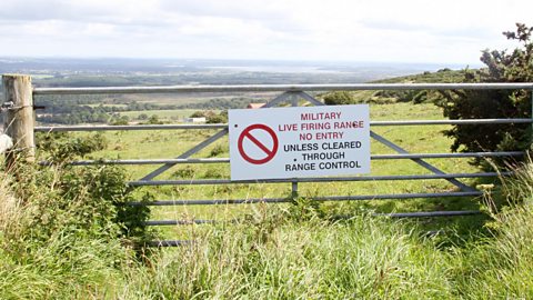 Sign on a field gate with the message 'Military live firing range no entry, unless cleared through range control' against the backdrop of a large field.