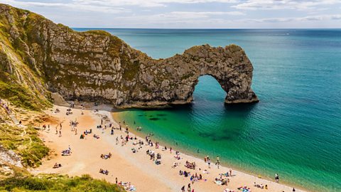 A beach which meets a large rock wall which extends into the sea where water passes through a large arch