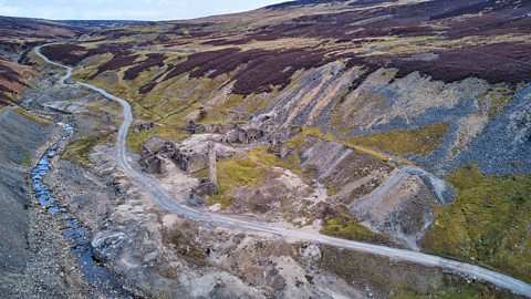 Aerial view of disused Old Gang lead smelting works, a road runs past ruined buildings in a steep and narrow valley surrounded with scree from industry.