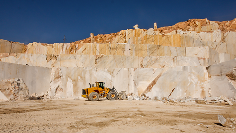 A marble quarry with large, stepped blocks of marble rock. The blocks' smooth surfaces have natural lines running through them. The rock is pale in colour, with shades of white, cream, and light orange.