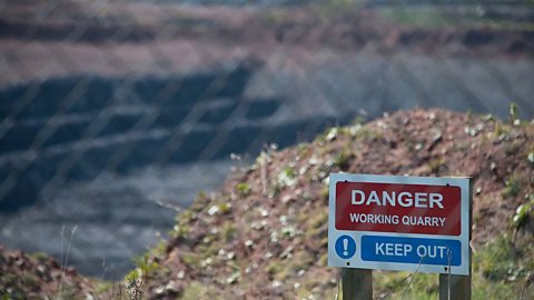 "Danger, Working quarry, Keep out!" sign with blurred quarry in the background and blurred fence in the foreground.