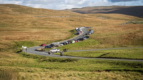 Cars are parked all over the road and outwith designated areas in a moor landscape