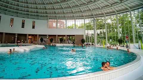 A large indoor thermal pool under a domed roof with people swimming or resting on the curved edge (Credit: Alamy)