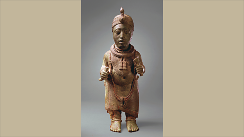 A small metal statue of a person wearing detailed decorations, including a necklace, patterned clothing, and a belt. The figure stands upright with both hands slightly raised.