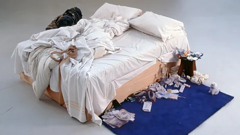 Tracey Emin's My Bed (1998) an unmade bed on a blue rug. The bed has white sheets and a pair of tights on the bed, with detritis gathered on the floor and on the bedside table (Credit: Courtesy of The Saatchi Gallery, London)