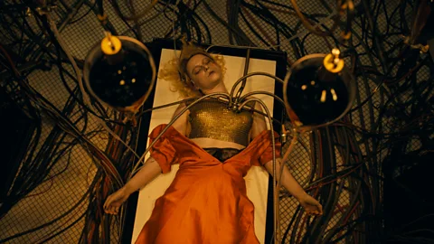 Jessie Buckley as The Bride with blonde hair, lying on table in red dress, with wires attached to her chest (Credit: Warner Bros)