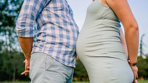 A man in a blue checked shirt touching bellies with a pregnant woman in a grey dress (Credit: Getty Images)