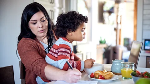 A mother holding a child on her lap as he eats breakfast and she writes on a paper (Credit: Getty Images)