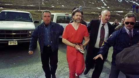 Theodore Kaczynski, the Unabomber, dressed in an orange prison jumpsuit and handcuffed, being led by federal agents (Credit: Getty Images)