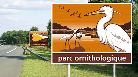 A road with a brown sign with three wading birds stood in water shown, and the words parc ornithologique at the bottom (Credit: Philippe Collier)