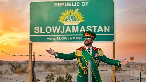 A bearded man in a green and gold military uniform with his arms outstretched behind a wire fence, with a billboard for the Republic of Slowjamastan behind him (Credit: Slowjamastan Ministry of Propaganda)