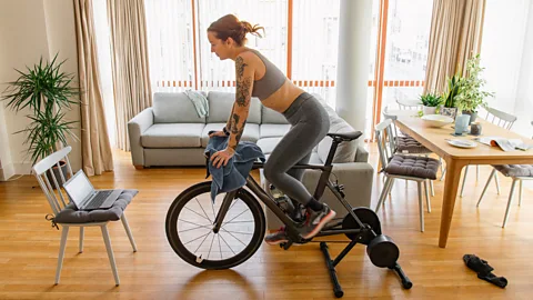 A woman pedalling on an exercise bike in her living room with a laptop on a chair in front of her (Credit: Getty Images)