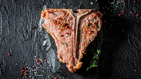 A thick T-bone steak sits against a black background, scattered with red peppercorns, rock salt and herb (Credit: Alamy)s
