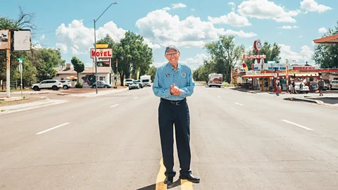 Angel Delgadillo stands in the middle of wide street on Route 66 in a postcard-style image reading "Greetings from Route 66" (Credit: Johnny Kompar)