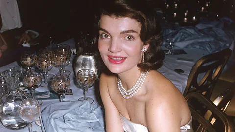 Jacqueline Kennedy Onassis poses for a photo sitting in a white dress and pearl necklace, with wine glasses on a white-tablecloth table behind her (Credit: Getty Images)