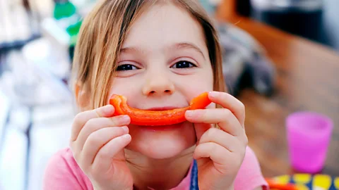 A young girl with blond hair holding a slice of red pepper in front of her mouth so it looks like a smile (Credit: Getty Images)