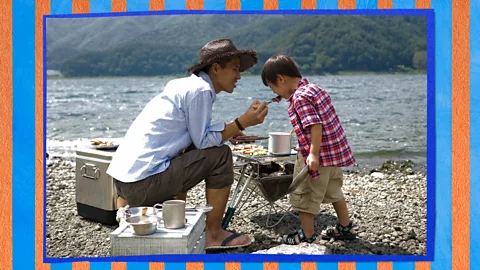 A father sitting down and feeding his young son along a lake, with a blue and orange scrapbook texture behind them (Credit: Serenity Strull/ BBC/ Getty Images)