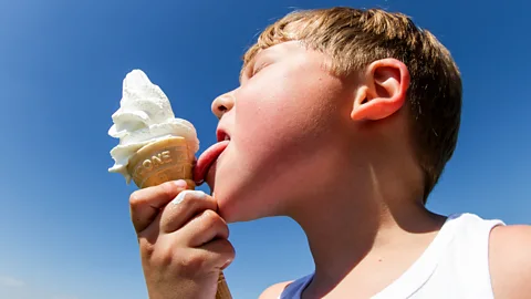 Young young boy in a white vest licks a soft srve vanilla icecream against the backdrop of a bright blue sky. (Credit: Getty Images)
