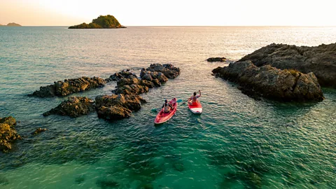 Two people paddle red kayaks through calm turquoise water among rocky outcrops at sunset (Credit: Getty Images)