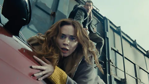 Josh O'Connor climbs a shipping container while Emily Blunt hangs on to the side of a red car in Disclosure Day (Credit: Universal Pictures)