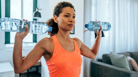 A woman in an orange sports top with a water bottle in each hand performing a shoulder press (Credit: Getty Images)