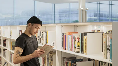 Person with a black beanie and a black t-shirt reads a book while standing in front of a library shelf. (Credit: Oodi Library, Helsinki)