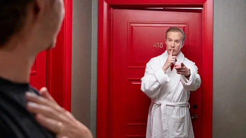 William Hanson wears white dressing gown and shushes someone in a hotel corridor (Credit: Olly Courtney for Hotels.com)