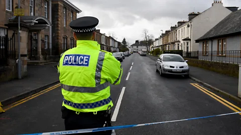 A police officer in a high-visibility jacket standing behind blue and white police tape closing off a residential street in Glasgow (Credit: Getty Images)