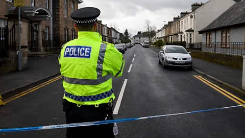 A police officer in a high-visibility jacket standing behind blue and white police tape closing off a residential street in Glasgow (Credit: Getty Images)