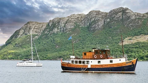 A small cruise boat and a yaught in front of a craggy and green hill (Credit: Alamy)