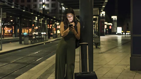 A woman stands beside a suitcase at a tram stop at night while looking at her mobile phone (Credit: Getty Images)